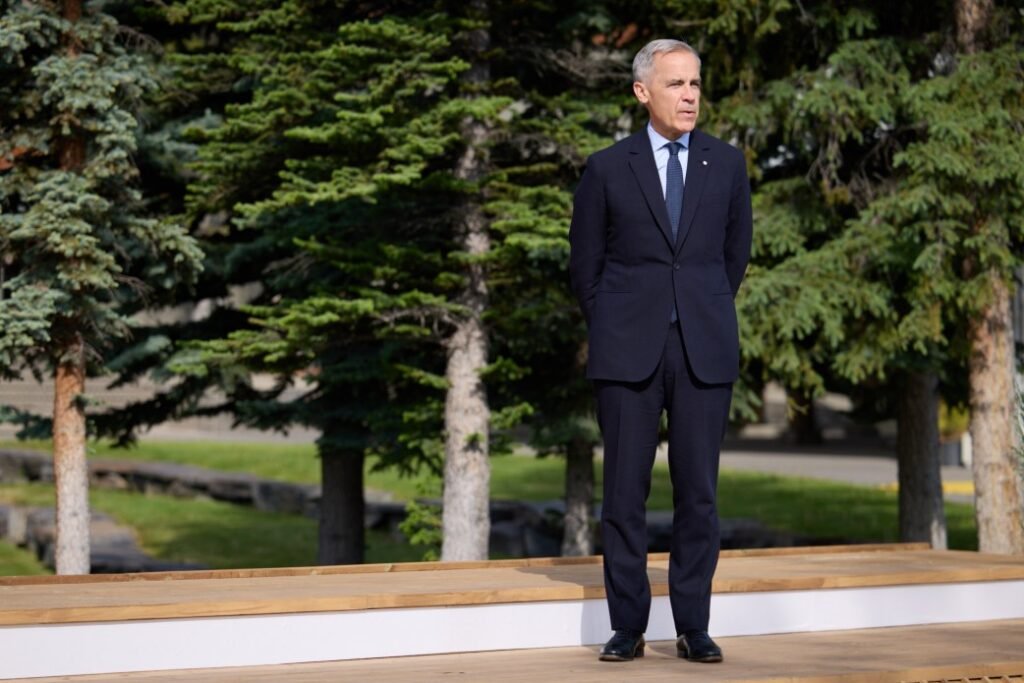 Марк Карні, чоловік у темно-синьому костюмі та синій краватці, стоїть на дерев'яному подіумі на відкритому повітрі. На задньому плані видно зелені дерева та будівлю.