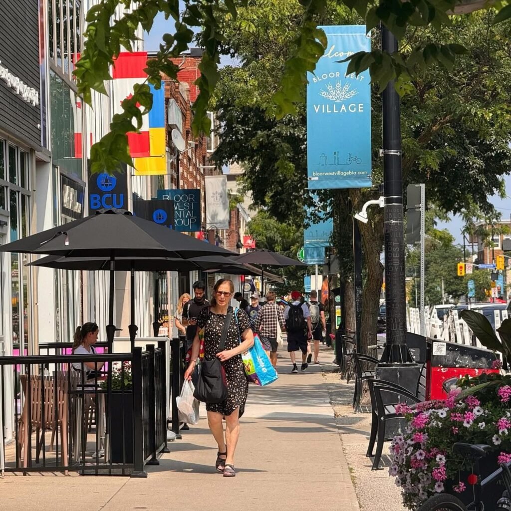 Сонячна вулиця в районі Bloor West Village з перехожими, вуличним кафе під чорними парасольками та блакитним банером району. На задньому плані вивіска BCU Financial.