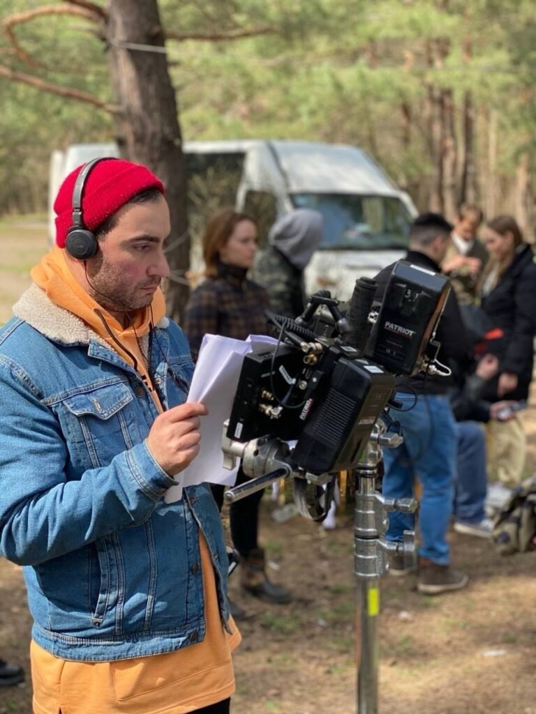 Режисер Гованес Хачатрян у червоній шапці та навушниках вивчає сценарій біля кінокамери на знімальному майданчику в лісі.