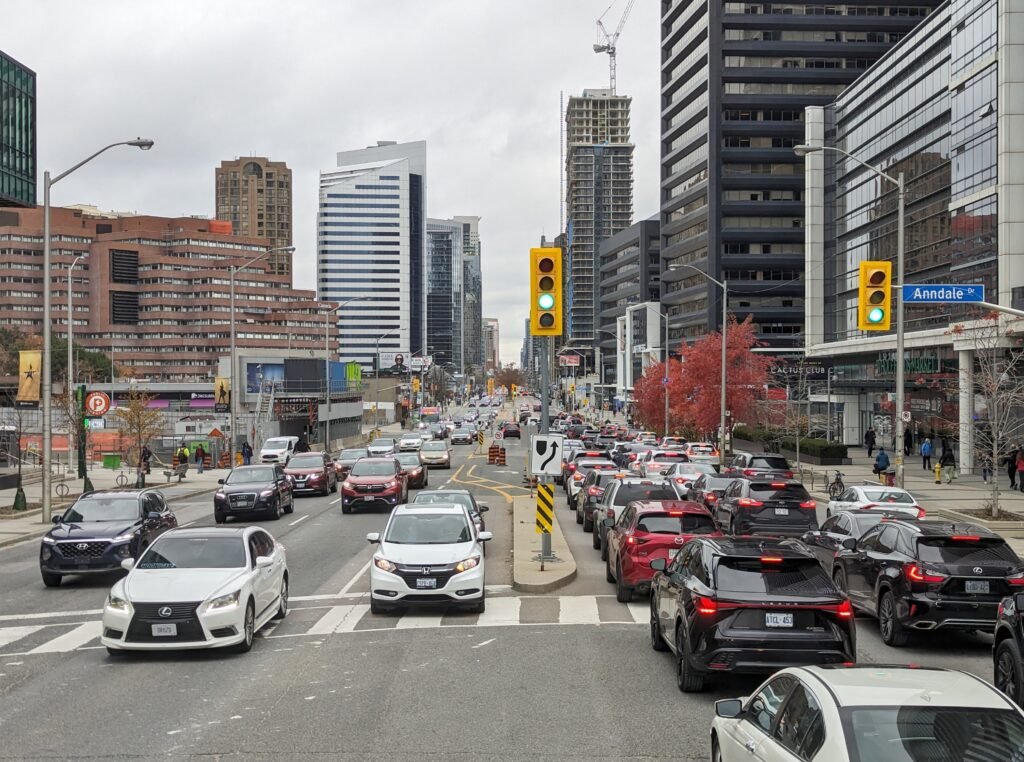 Жвавий рух автомобілів на перехресті вулиць Yonge Street та Anndale Drive у районі North York, Торонто, оточеному хмарочосами та офісними будівлями під похмурим небом.