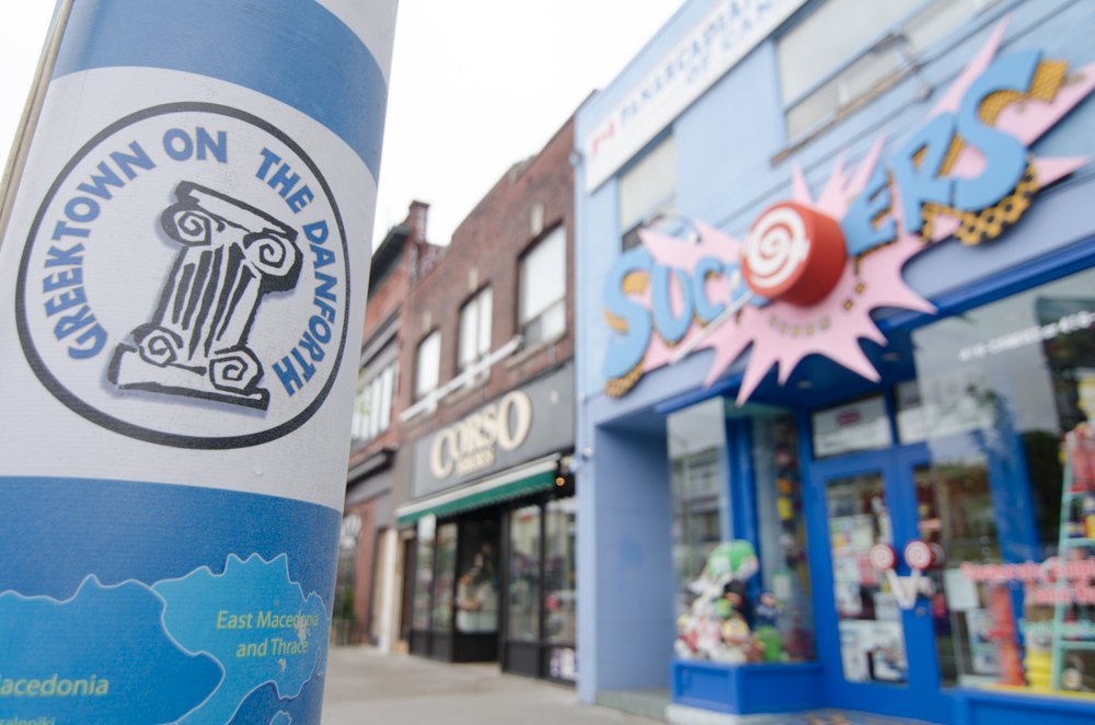 Інформаційна наклейка з логотипом Greektown on the Danforth на стовпі на фоні магазинів у Торонто.