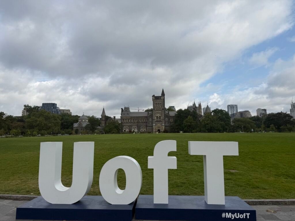 Великі білі літери UofT на передньому плані на фоні зеленого газону та історичної кам'яної будівлі кампусу St. George Університету Торонто.