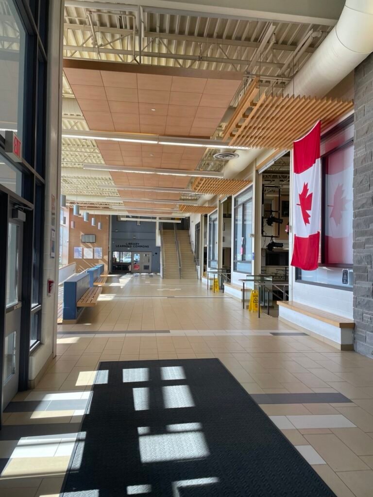 Світле та просторе лобі канадської старшої школи Garth Webb Secondary School із прапором Канади на стіні, сучасним дизайном стелі та входом до бібліотеки.