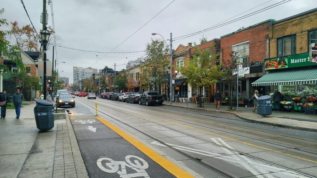 Вулиця Roncesvalles Avenue у Торонто з трамвайними коліями, велодоріжкою та невеликими крамницями під хмарним небом.