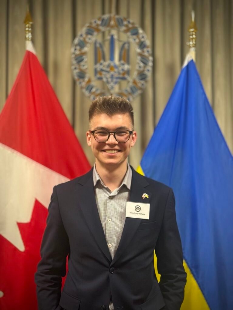 Молодий чоловік у темному піджаку та окулярах з бейджем Volodymyr Seheda посміхається на фоні прапорів Канади та України.