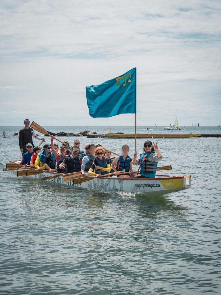 Група людей у великому човні-драконі на воді тримає весла та підіймає блакитний кримськотатарський прапор із золотою тамгою.