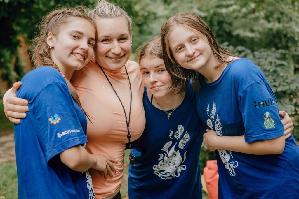 Засновниця проєкту Оксана Грицина обіймає трьох дівчат-підлітків у синіх брендованих футболках табору Соняшник на фоні природи.