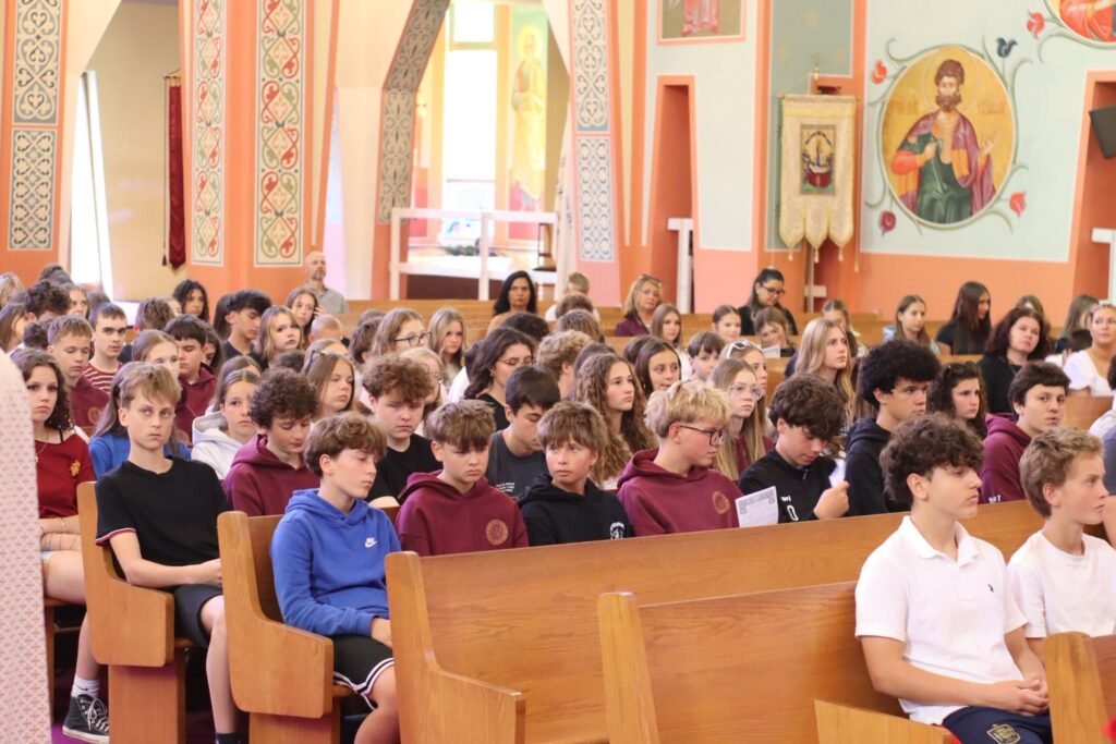 Велика група підлітків, учнів 8-х класів українських шкіл, сидять на дерев'яних лавах у храмі під час урочистого заходу.