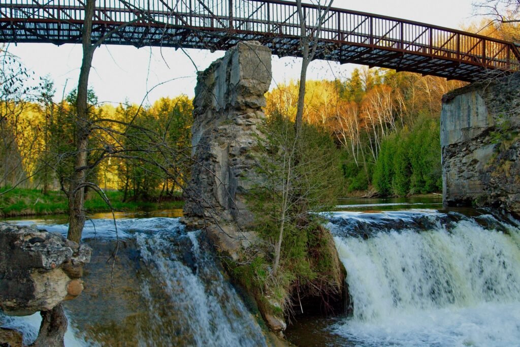 Водоспад у парку Forks of the Credit з кам'яною руїною в центрі та металевим пішохідним мостом зверху на фоні осіннього лісу.