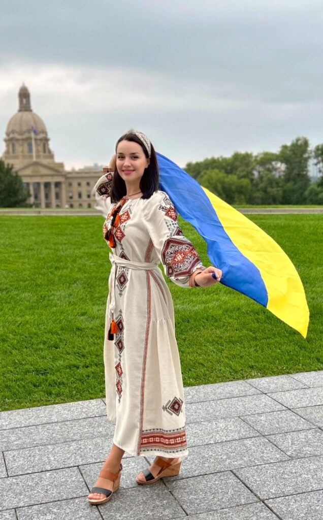 Жінка у довгій білій вишитій сукні тримає розгорнутий синьо-жовтий прапор України на фоні будівлі законодавчих зборів провінції Альберта в Едмонтоні.