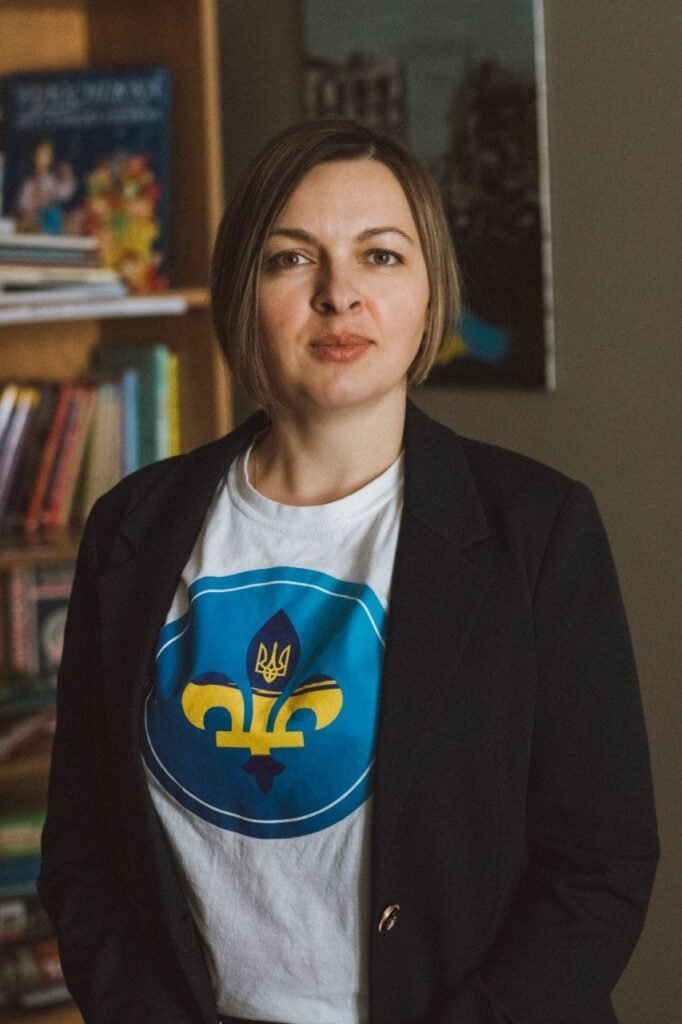Портрет Богдани Поради у білій футболці з логотипом, що поєднує український тризуб та квебекську лілію, у синьому піджаку на фоні книг.