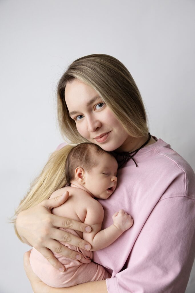 Світлошкіра жінка з довгим білявим волоссям у рожевому світшоті ніжно тримає на руках спляче немовля на світлому фоні.