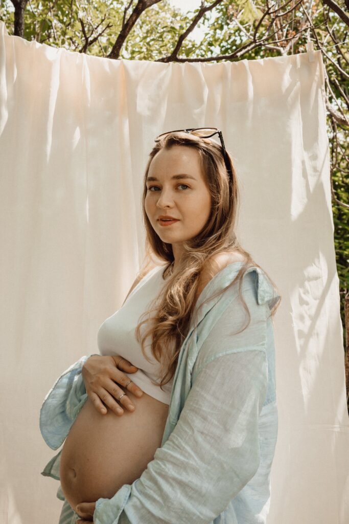 Вагітна Iryna Litva у блакитній сорочці тримає руки на животі на фоні природи
