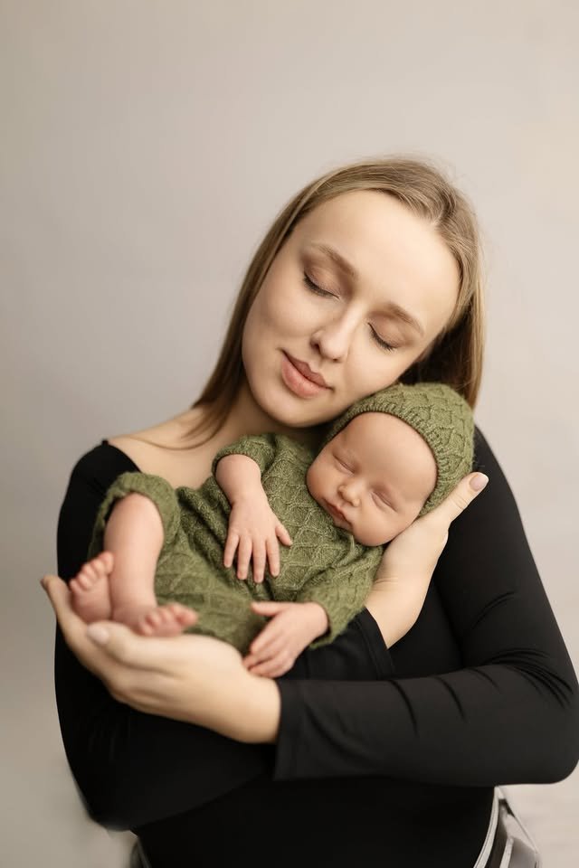 Жінка з довгим світлим волоссям обіймає спляче немовля у зеленому в'язаному костюмі.