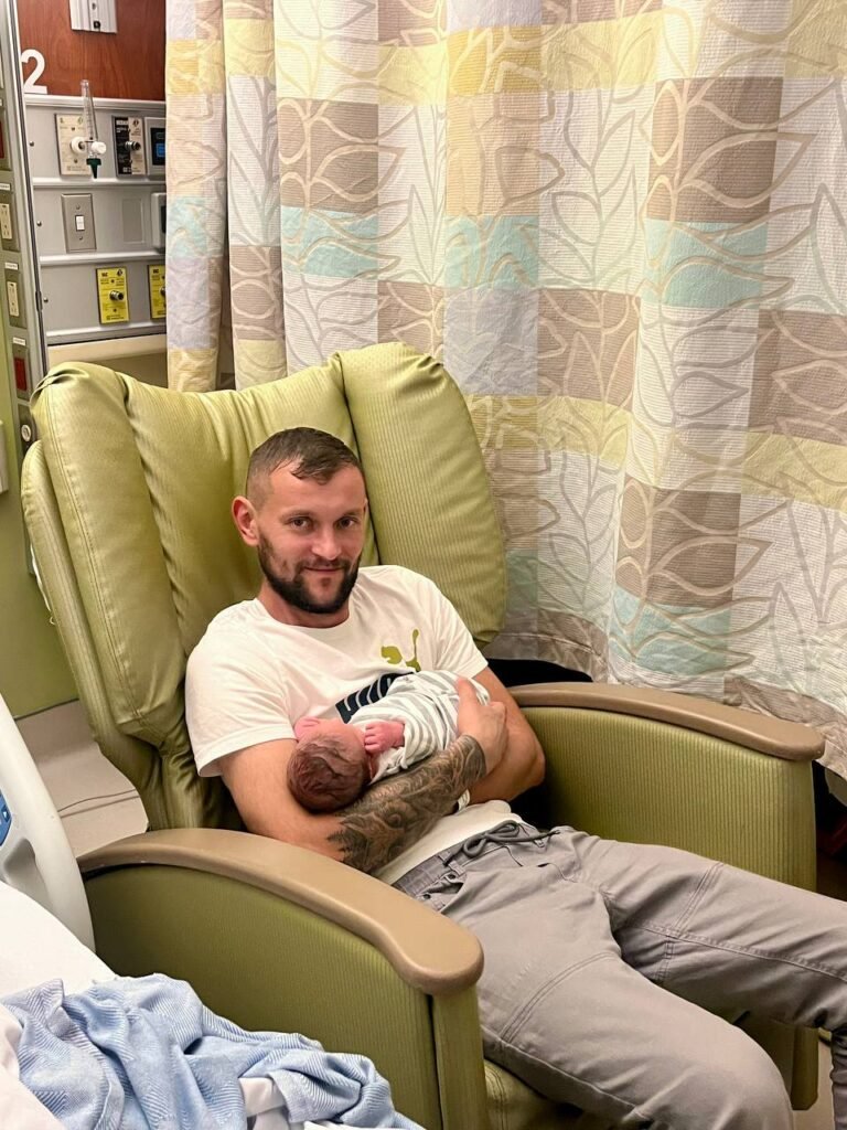 Чоловік у лікарняній палаті тримає на руках новонароджене немовля, загорнуте в смугасту пелюшку
