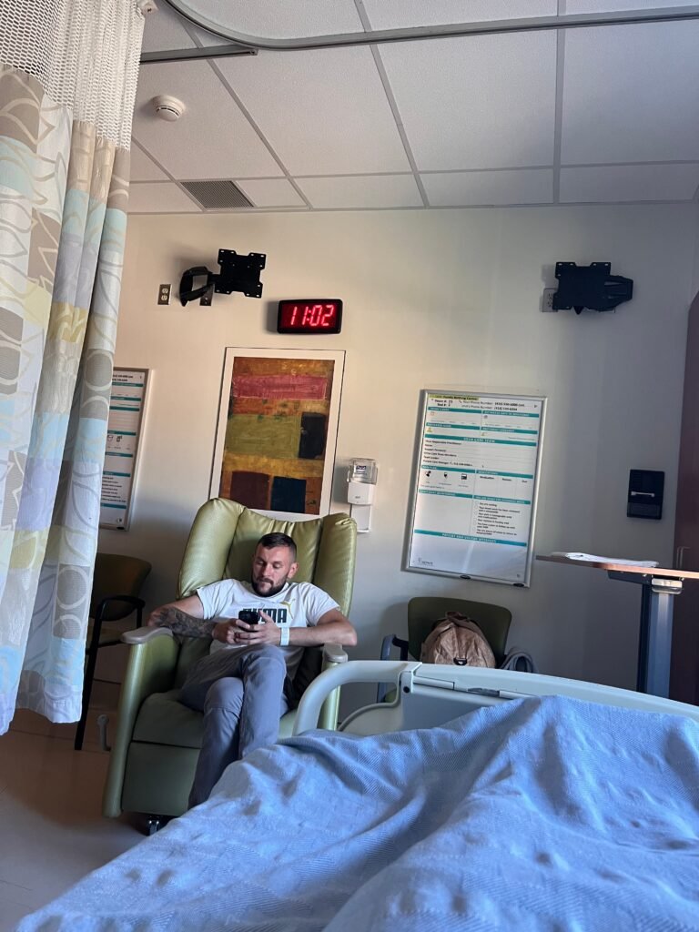 Чоловік сидить у медичному кріслі в сучасній лікарняній палаті в Канаді біля ліжка породіллі.