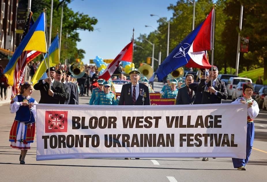 Учасники параду в Торонто несуть великий білий банер з написом Bloor West Village Toronto Ukrainian Festival. На фоні видно прапори України, Канади та НАТО, дітей у народних строях та чоловіків у військовій формі.