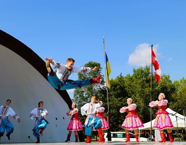 Група танцюристів в українських народних костюмах на сцені просто неба, центральний виконавець у високому стрибку на фоні прапорів України та Канади.