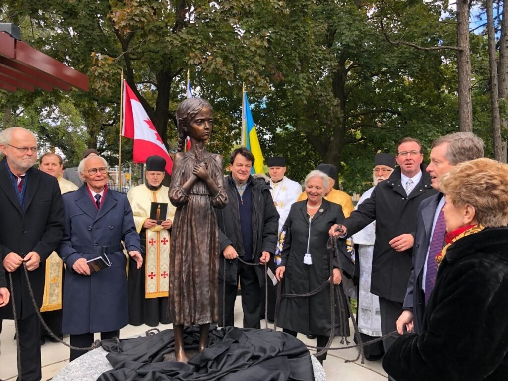 Бронзова статуя дівчинки з колосками пшениці під час церемонії відкриття меморіалу жертвам Голодомору в Торонто на фоні прапорів України та Канади.