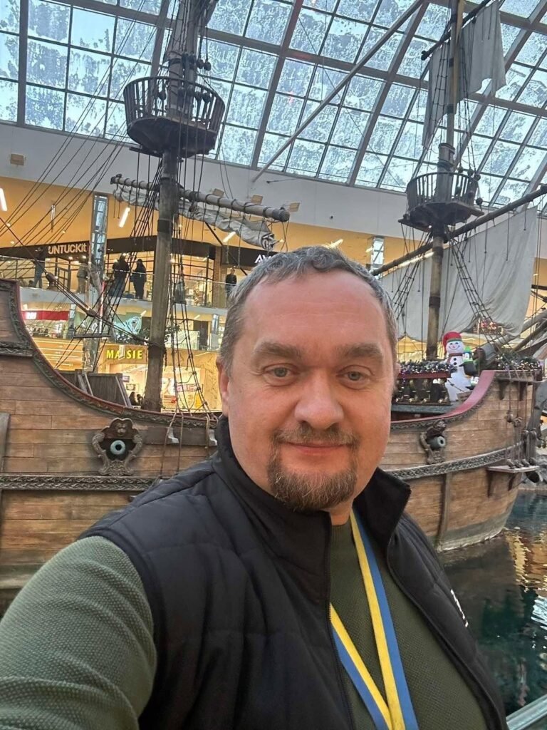 Селфі чоловіка на ім'я Vitaliy Kushnir у чорній жилетці із синьо-жовтою стрічкою на шиї на тлі дерев'яного вітрильного корабля в атріумі торгового центру.