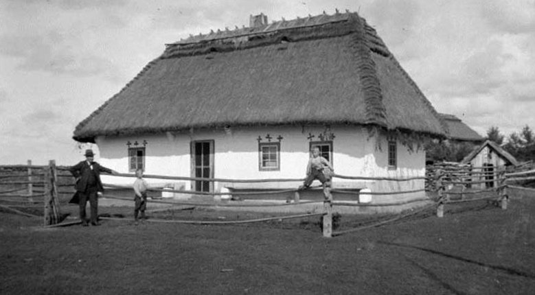 Чорно-біле історичне фото традиційної української біленої хати під солом'яною стріхою в Манітобі. Перед будинком стоять чоловік у капелюсі та двоє дітей біля дерев'яної огорожі.