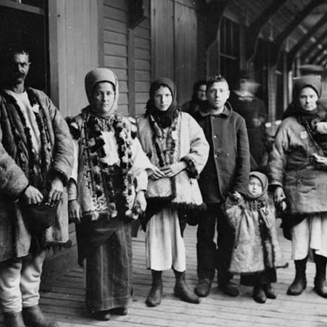 Чорно-біле архівне фото української родини в традиційних кожухах та хустках на дерев'яному пероні в Канаді.