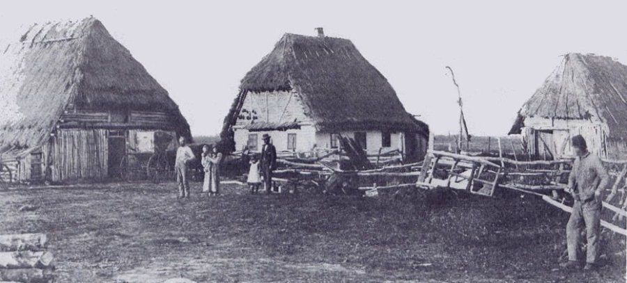 Історичне чорно-біле фото трьох традиційних українських хат під стріхою та родини переселенців у канадських преріях на початку XX століття.