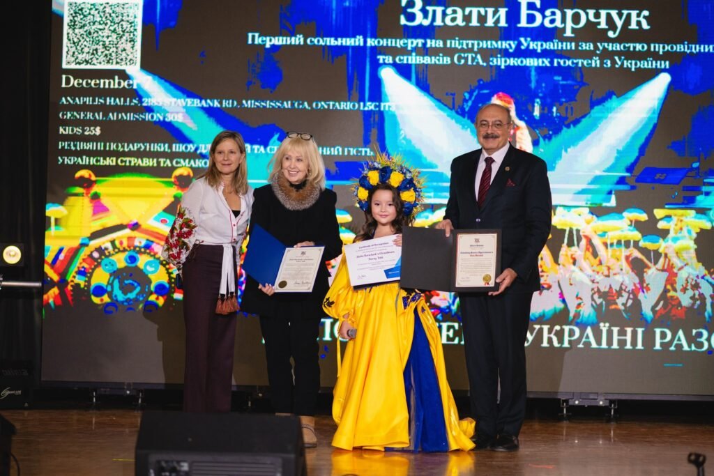 Злата Барчук у жовто-синій сценічній сукні та вінку тримає грамоту на сцені у супроводі трьох дорослих під час нагородження.