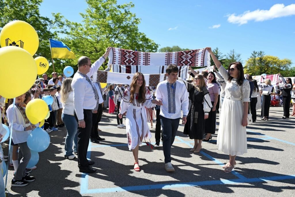 Учні та дорослі у вишиванках тримають вишиті рушники над випускниками під час урочистої церемонії на відкритому повітрі.
