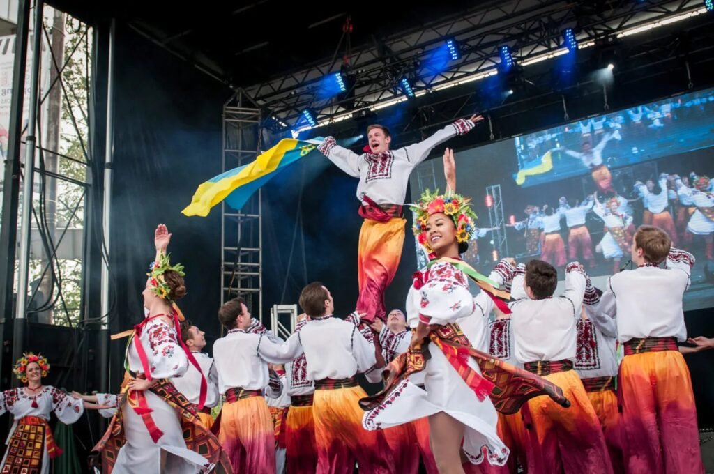 Танцюристи в українських народних костюмах виконують складний хореографічний номер на сцені, один з артистів піднятий угору з українським прапором.