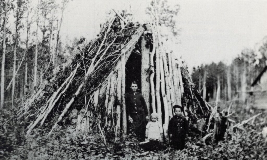 Історичне чорно-біле фото української родини переселенців — жінки та двох дітей — біля примітивного тимчасового укриття з колод і гілок у лісистій місцевості Канади.