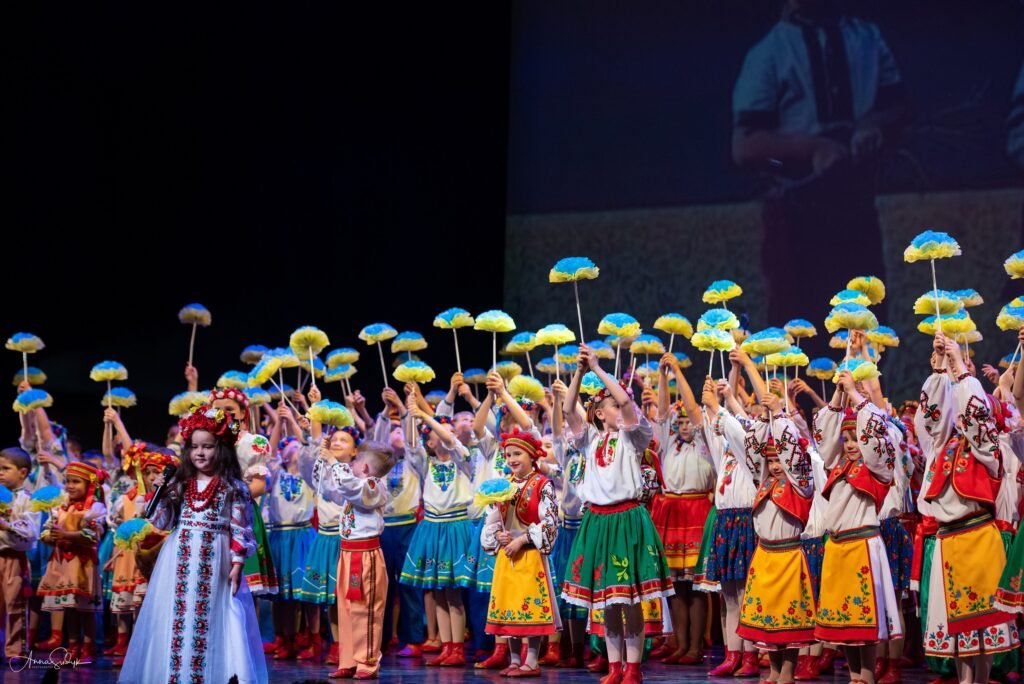 Юна співачка Злата Барчук у білій вишитій сукні та червоному вінку співає на сцені перед великим дитячим колективом у народних строях із синьо-жовтими квітами.