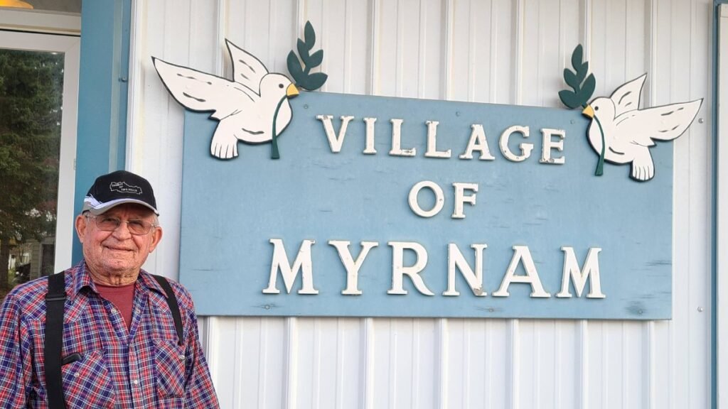 Літній чоловік у картатій сорочці стоїть поруч із вивіскою Village of Myrnam, прикрашеною символічними білими голубами миру.