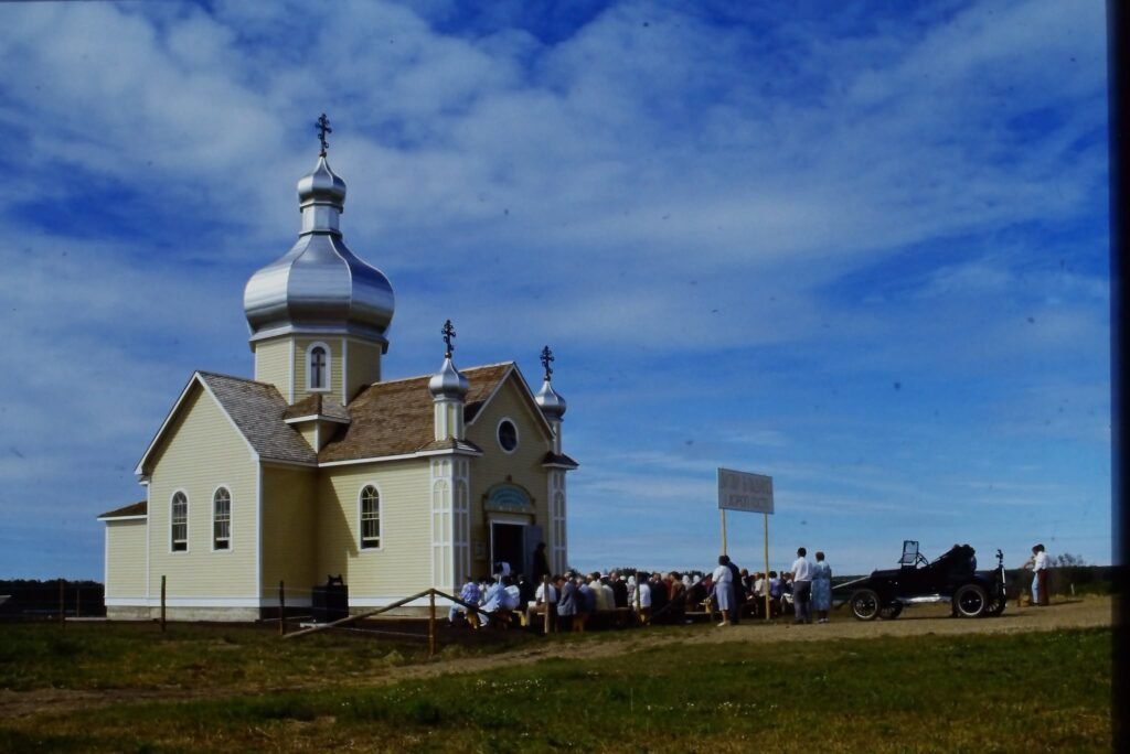 Жовта дерев'яна церква зі срібними куполами, перед якою зібралися люди на церемонію освячення в канадському музеї просто неба.